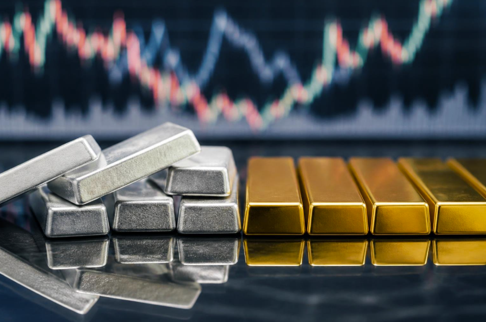 Stacks of silver bars and gold ingots on a reflective surface with a blurred stock chart in the background, conveying precious metals trading.
